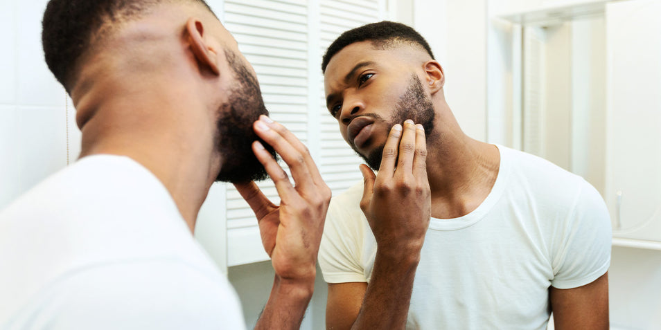 A man touching his thinning beard in front of a mirror